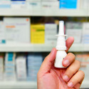 Photo of a hand holding a bottle of nasal spray against a pharmacy's stock shelves.