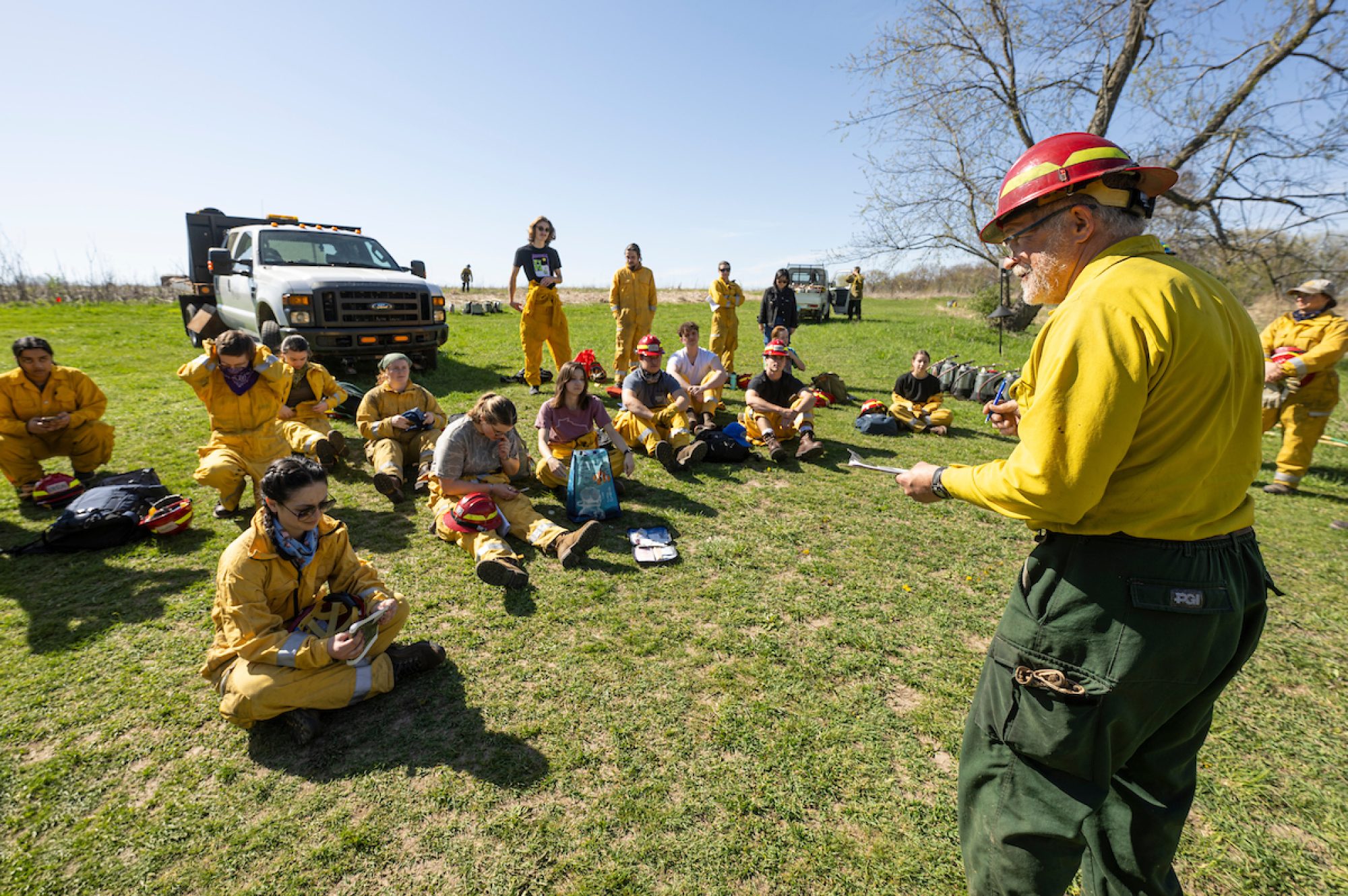 A group of students sit outside on the grass in front of a work truck, facing an older man standing before them who appears to be giving them instructions. Students are wearing bright yellow long-sleeved shirt, some with matching pants, and have red hard hats, note books and personal belongings beside them on the grass. The older man also wears a bright yellow long-sleeved shirt and his red hard hat with dark green pants.