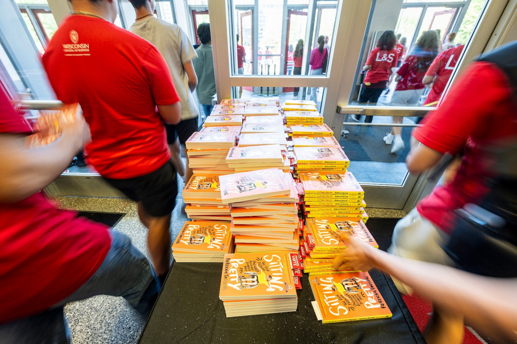 A bunch of books sit on a table as students walk by and grab them.