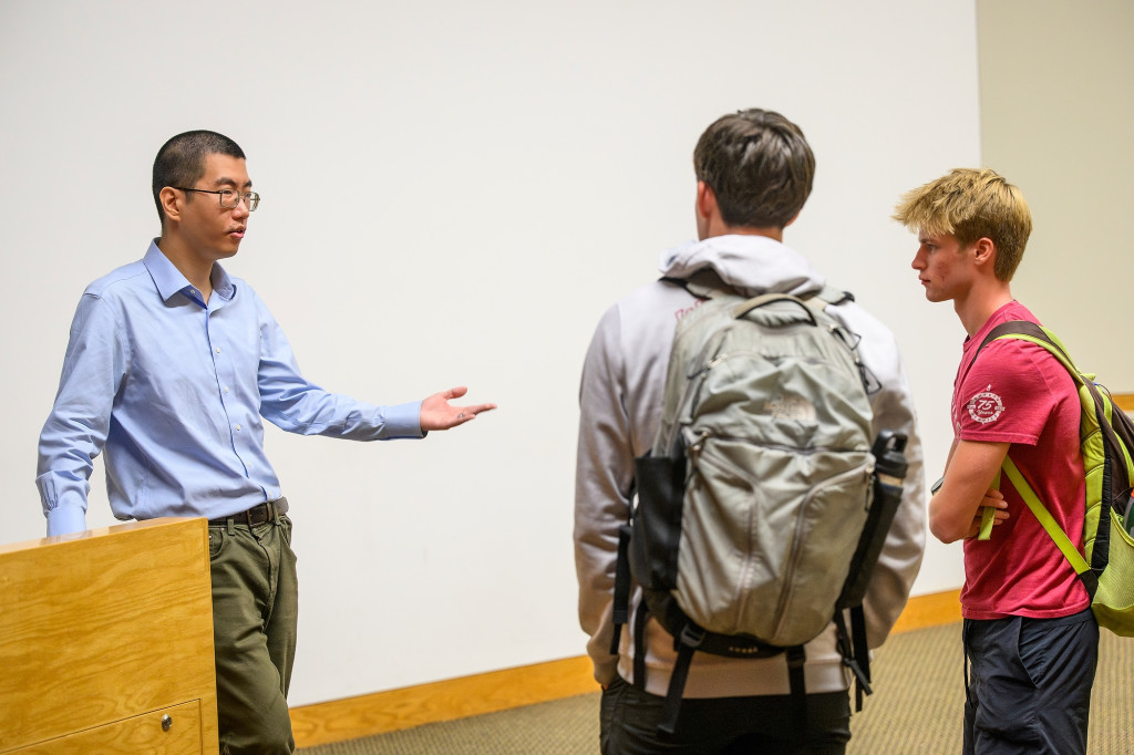 A professor gestures as he talks to two students wearing backpacks.