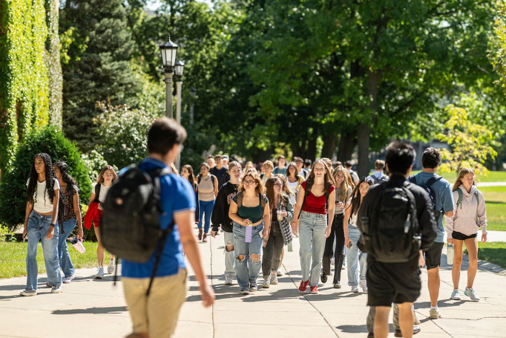 Several young men and women with backpacks are walking along a sidewalk surrounded by trees.