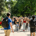 Several young men and women carrying backpacks walk up a tree-lined sidewalk.