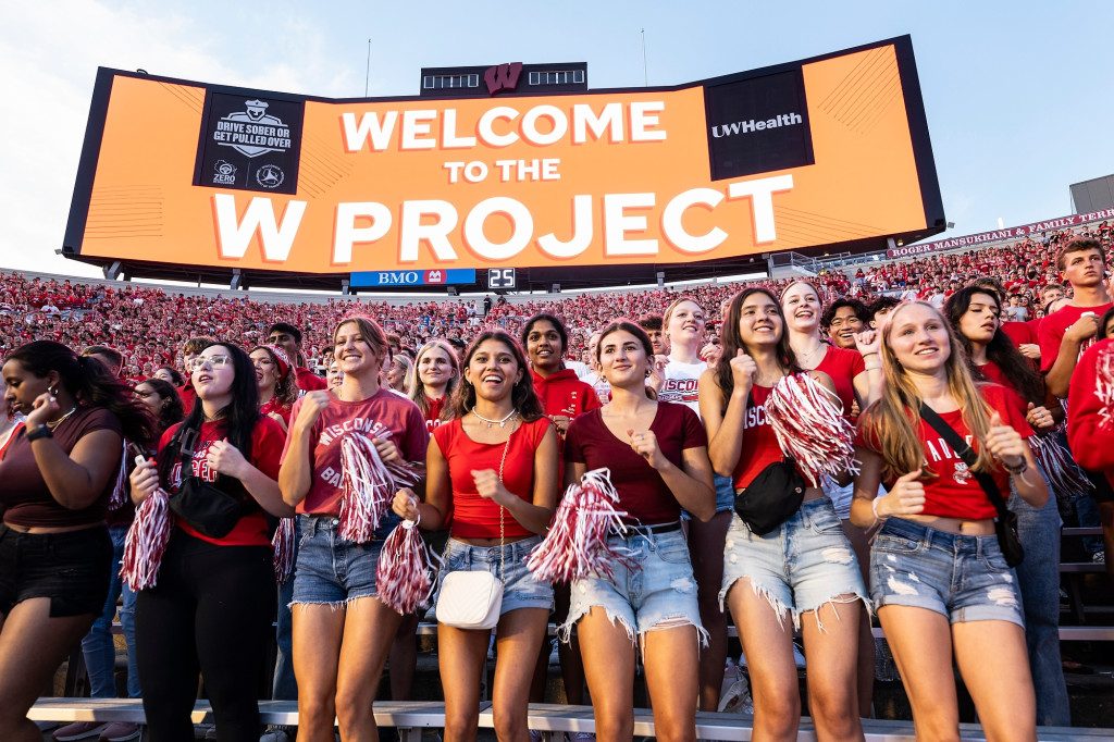 A group of first-year students stand in the bleachers at Camp Randall Stadium, cheering and smiling.