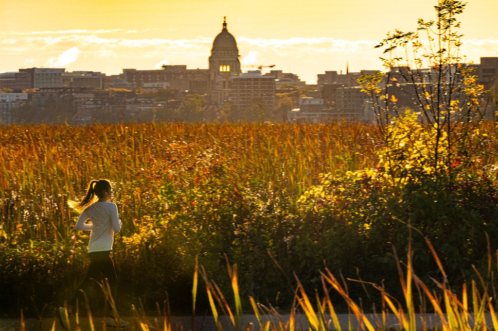 A jogger is seen running on a path surrounded by trees with colorful leaves; the capitol building is in the background.