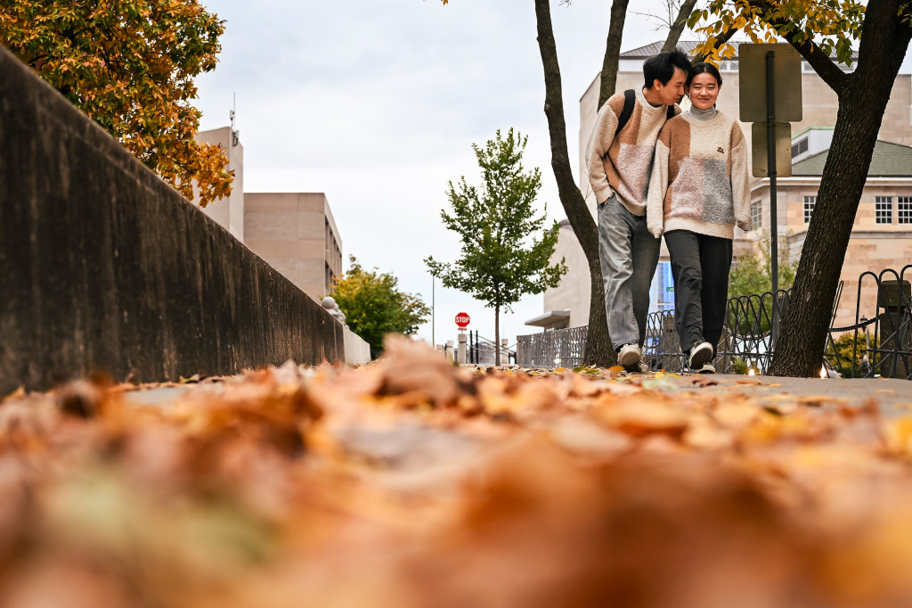 Two people huddle together and talk on a leaf-covered sidewalk.