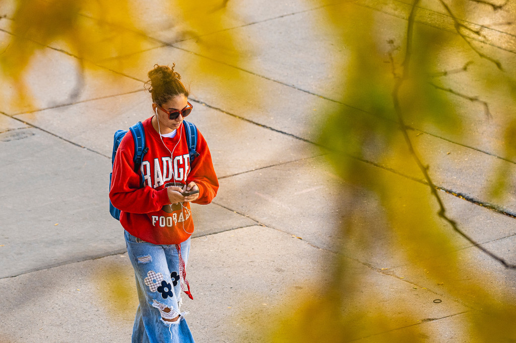 A woman wearing a Wisconsin sweatshirt is seen walking under tree branches covered with yellow and green leaves.