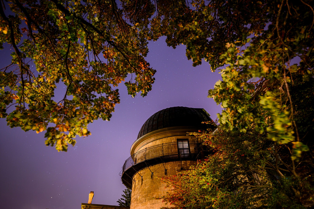 Northern lights are visible in the sky behind an observatory dome and an oak tree.