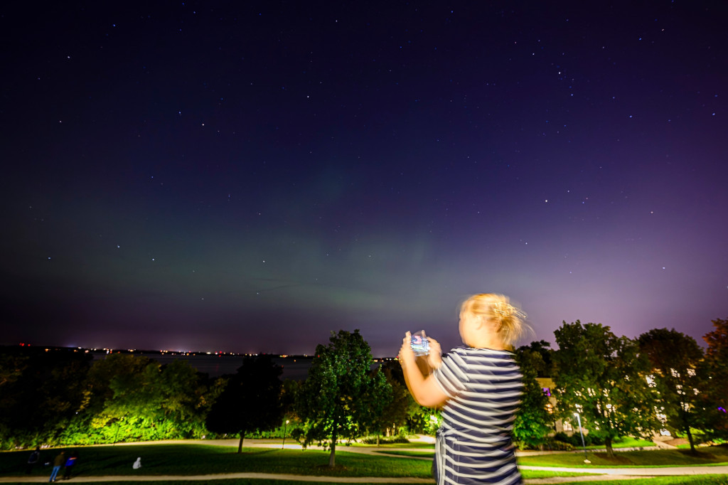 A woman in the foreground holds a phone to take pictures of lights in the nighttime sky, visible in the background.