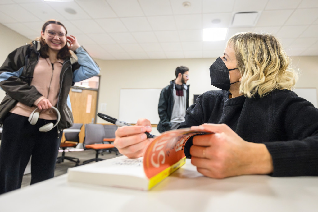 A woman holds a book and signs inside the cover while chatting with a student.