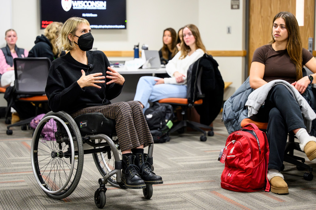 A woman in a wheelchair sits in the middle of a classroom and speaks, gesturing.