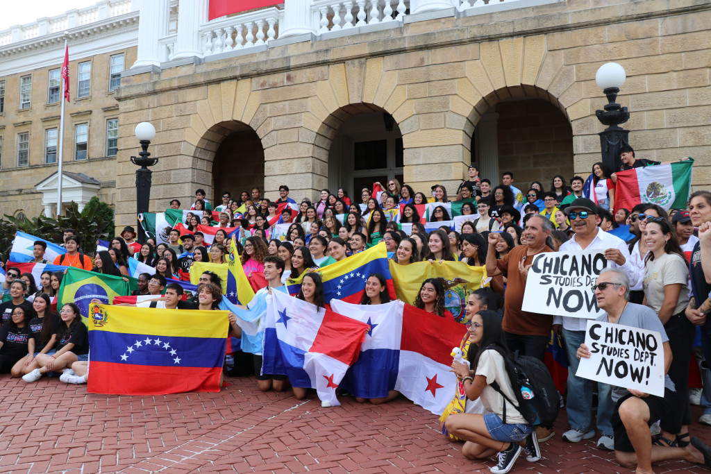 A large group holds Latine flags and signs while posing for a picture in front of a university building.