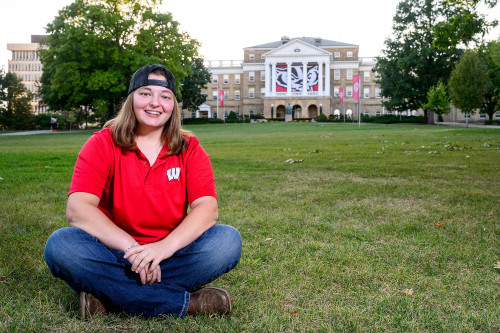 A woman in a red shirt and jeans sits on a lawn with a building behind her.