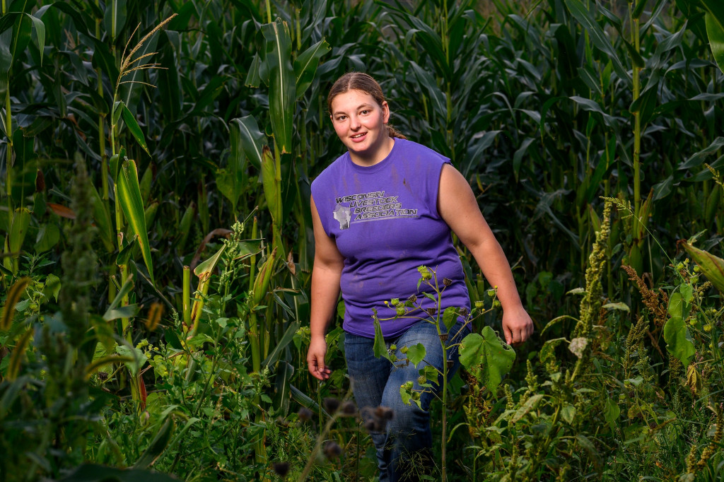 A woman walks through a cornfield. She wears a purple t-shirt.