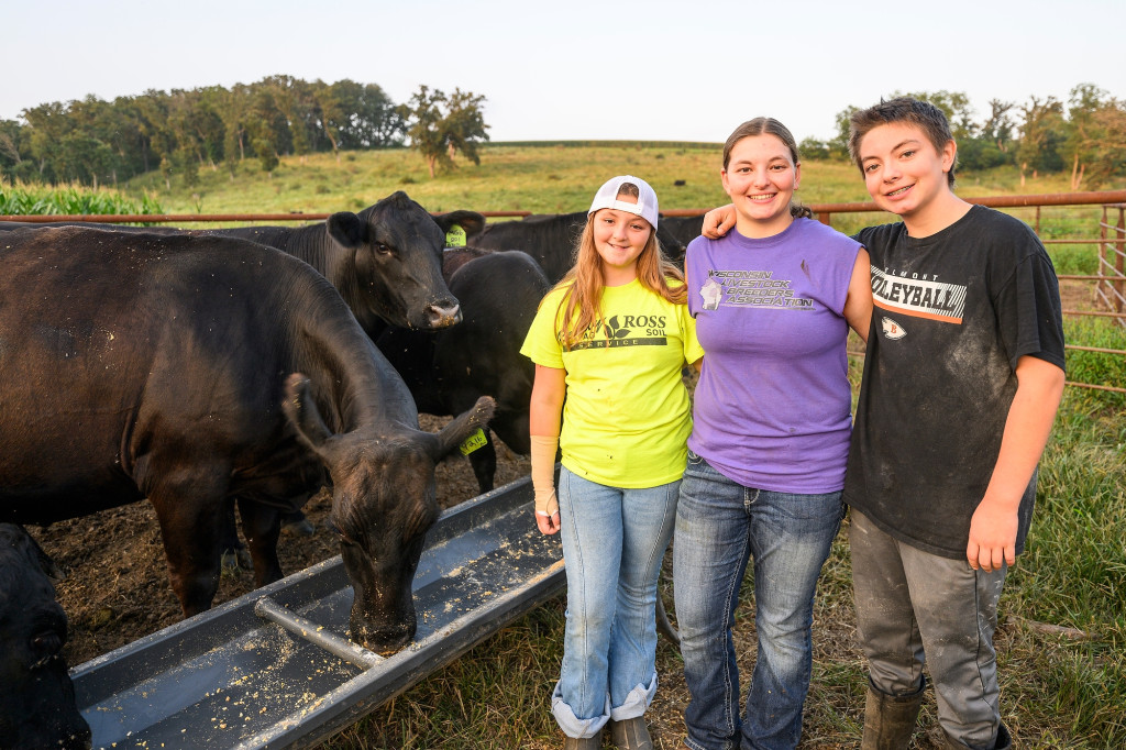 Three people wearing jeans and t shirts stand, arms around each other and smiling, as cows eat from a trough in the background.