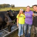 Three people wearing jeans and t shirts stand, arms around each other and smiling, as cows eat from a trough in the background.