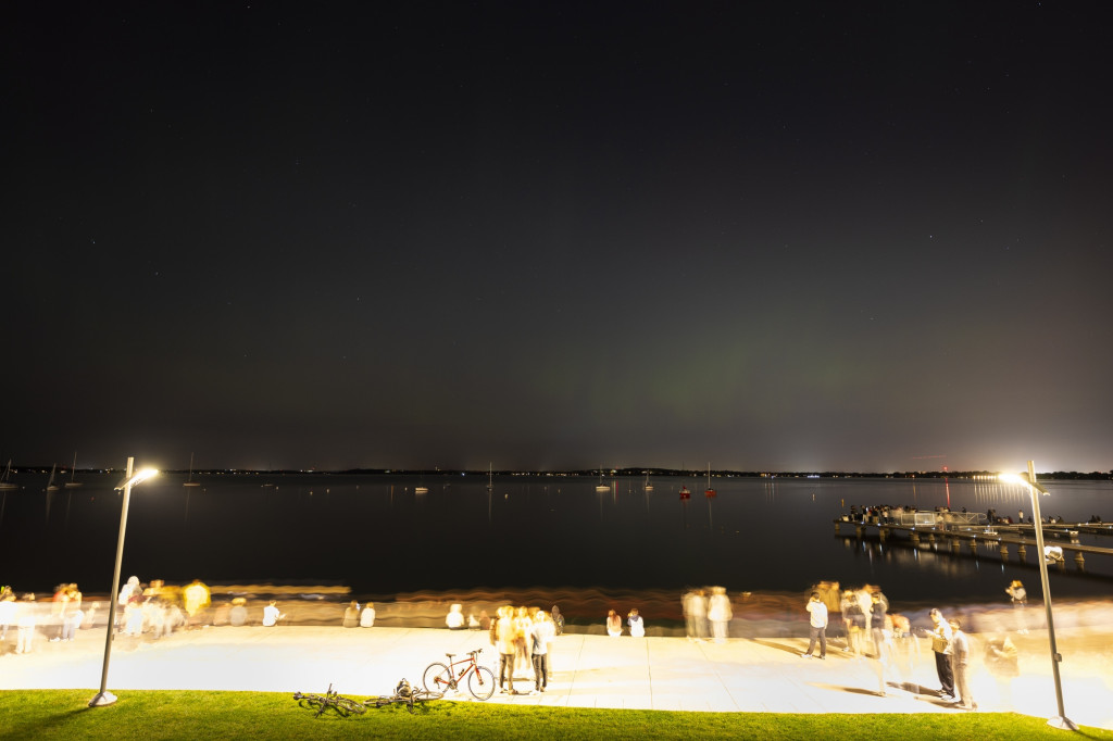 People stand on a well-lit terrace, with the dark night sky behind them. The sky is lit up by northern lights.