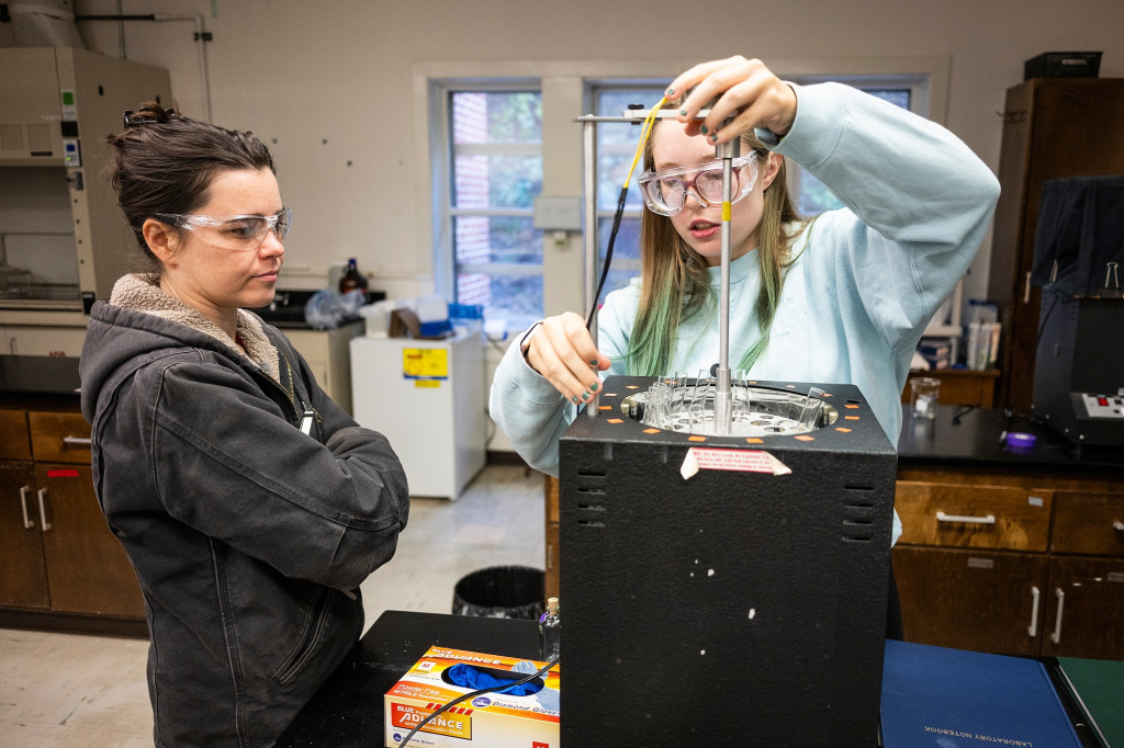 A woman works on lab equipment while another woman listens to her explain what she is doing.