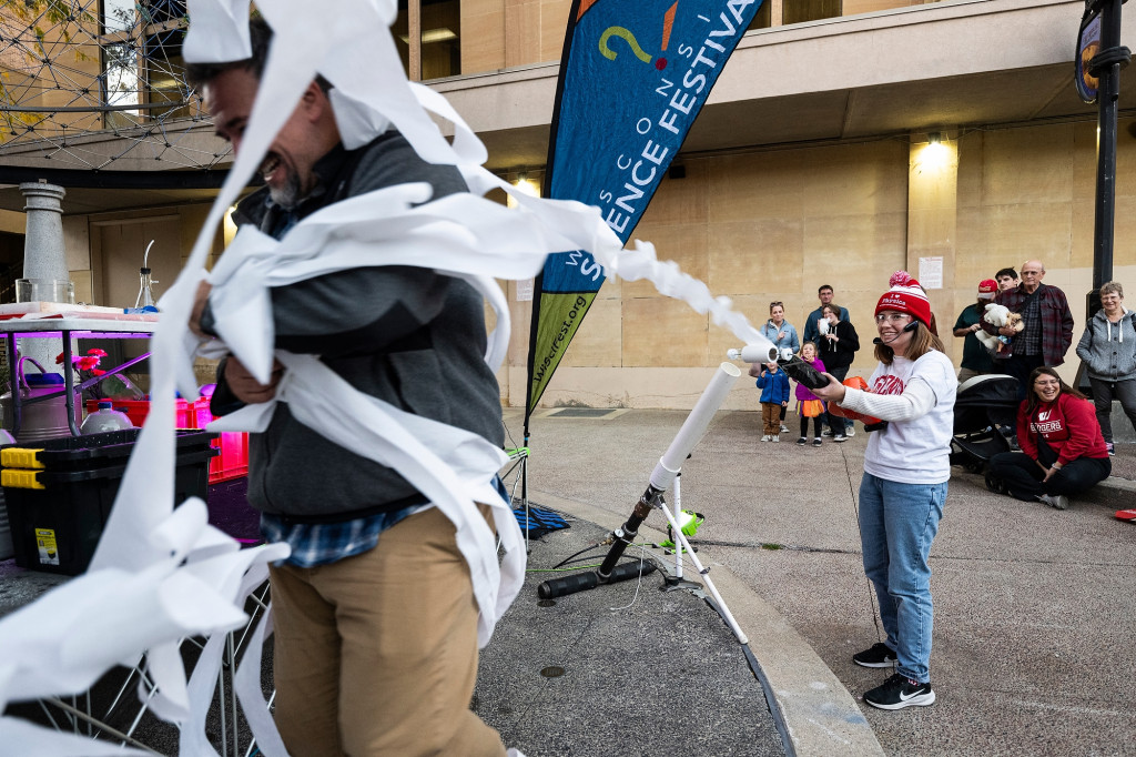 A woman blows toilet paper off a roll as another person is rolled up into it.