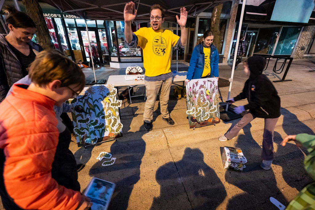 Two adults lead a game played by children crouching on the ground.