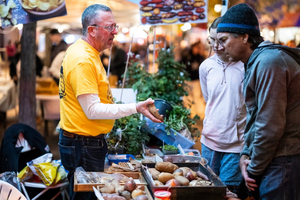 A man standing at a booth talks with people; sitting on a table nearby is a box of potatoes.