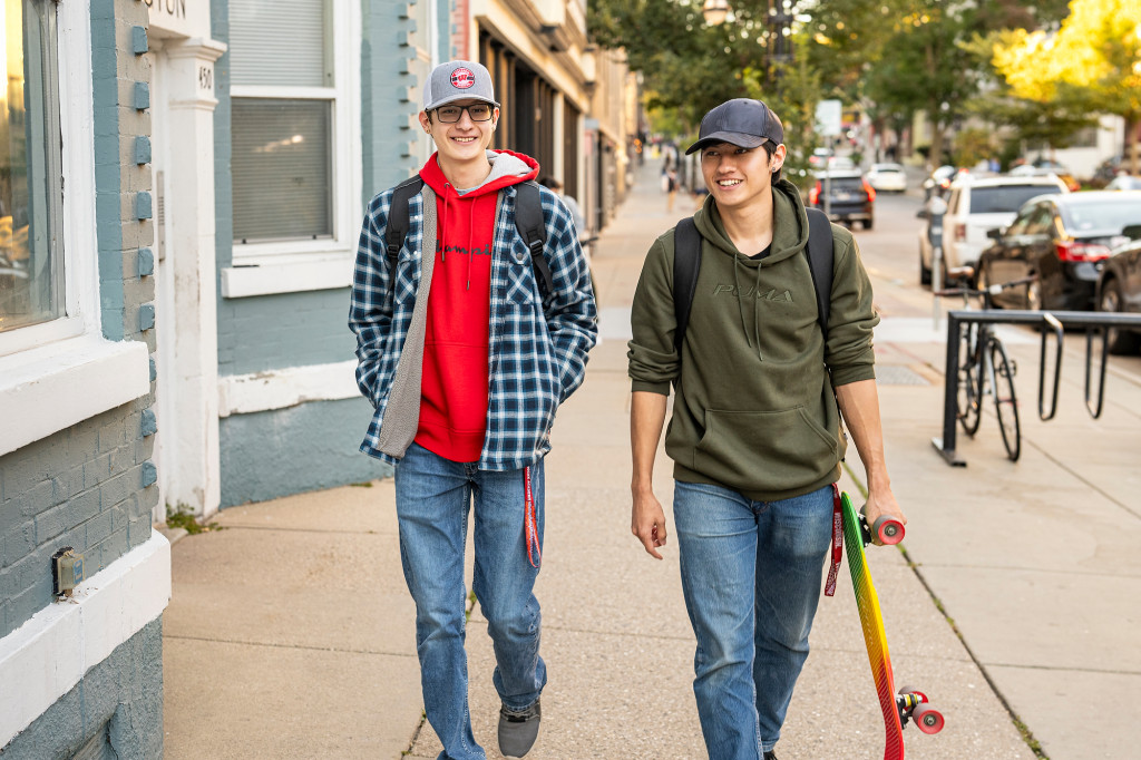 Two young men walk side by side, wearing informal clothes and talking. One carries a skateboard.