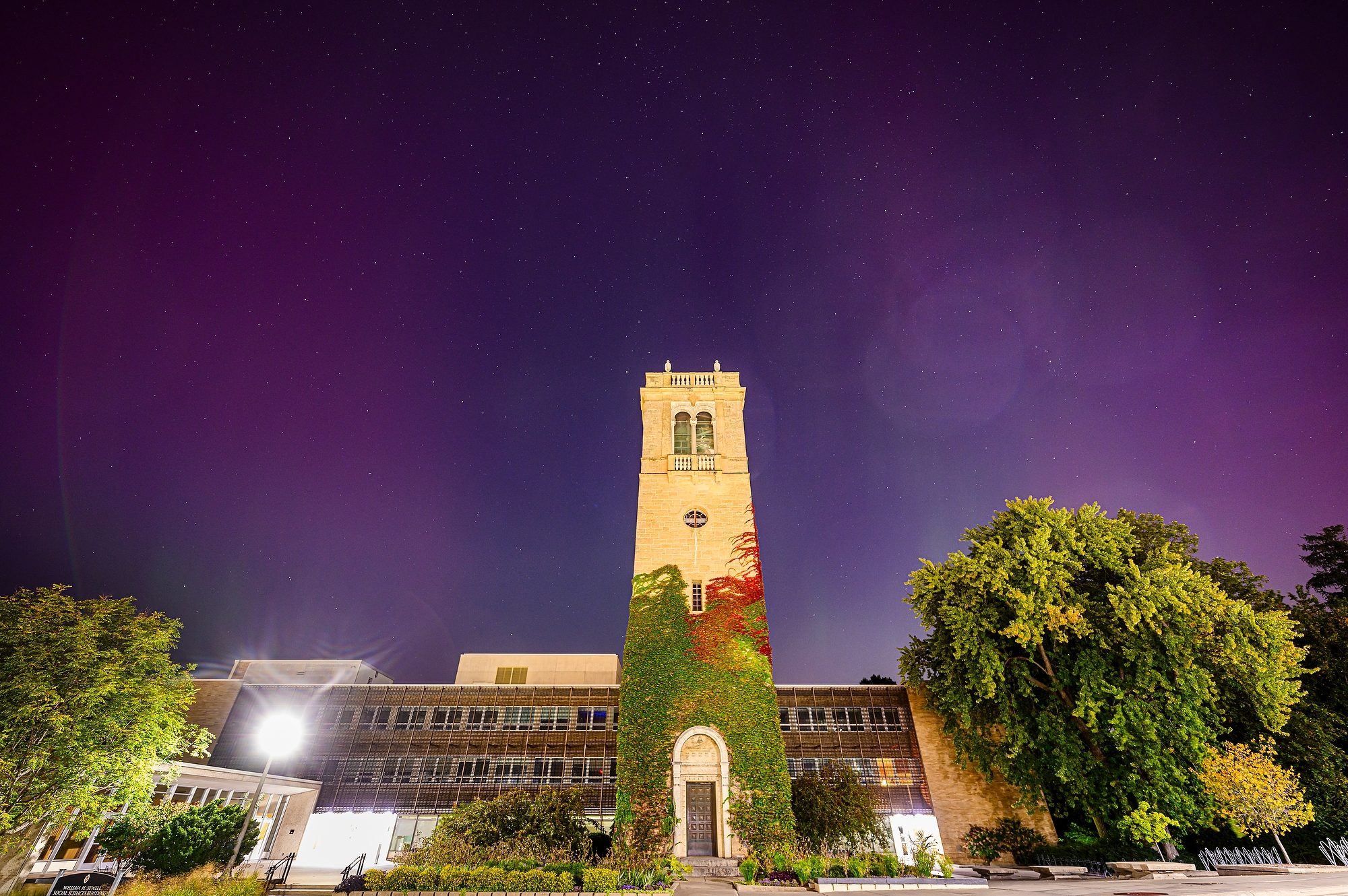 Night view of the iconic Carillon Tower at the University of Wisconsin-Madison, illuminated against a starry sky with vibrant purple hues of the aurora borealis.