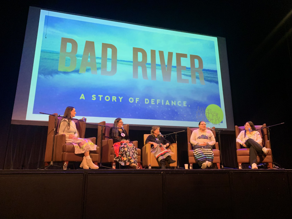 Five women sit on a stage in front of a large screen that reads, "Bad River A Story of Defiance."