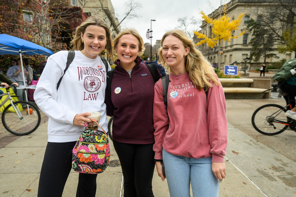 Three young women stand close together and smile for the camera. They wear 