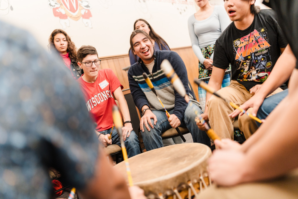 A group of Indigenous students are seated around a drum. They sing and smile while playing the drum.