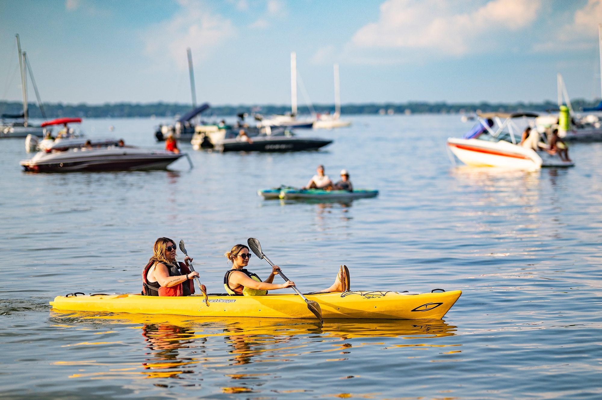 Two people kayaking in a yellow kayak float on Lake Mendota with several boats in the background on a sunny day.