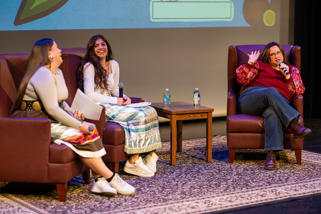 Two young women sit in chairs on a stage and laugh while a third women, also in a chair, makes a joke.