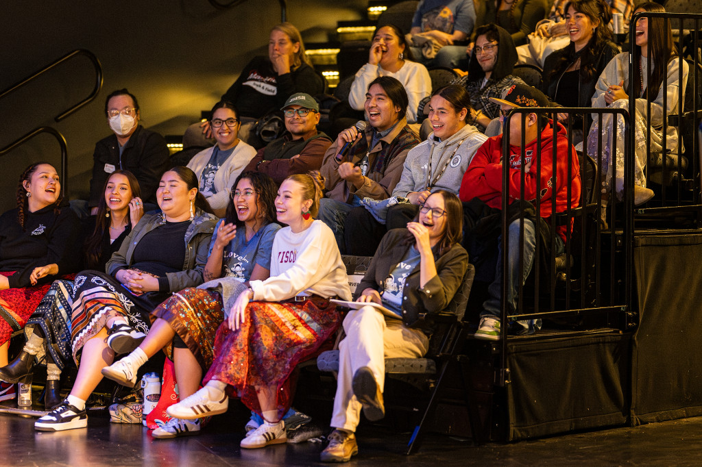 A crowd of students laugh at a joke while sitting in the audience of an indoor theater.