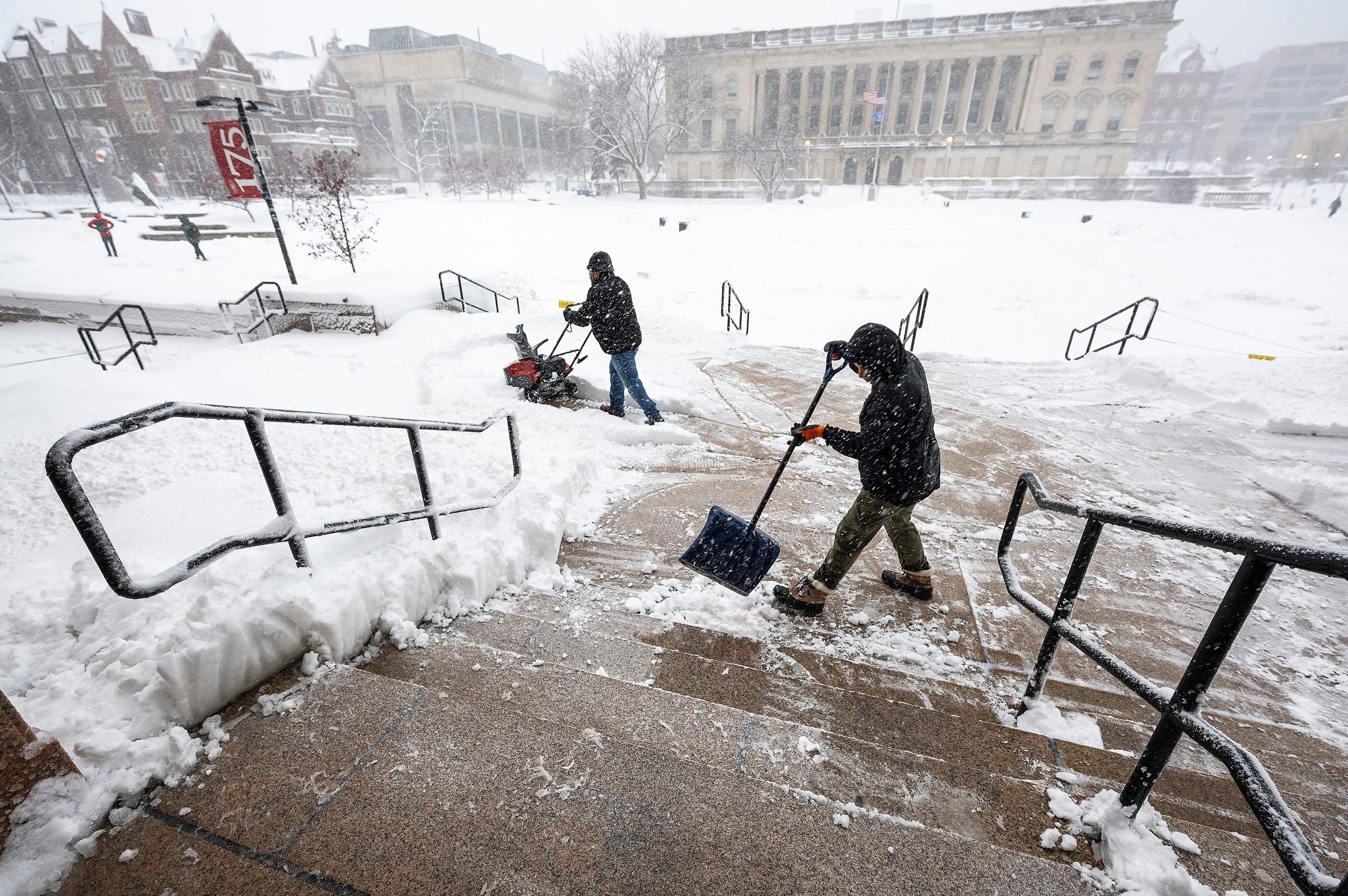 Two individuals clear snow from a pathway using a snow blower and a shovel on the UW–Madison campus, with snow-covered grounds and buildings visible in the background.