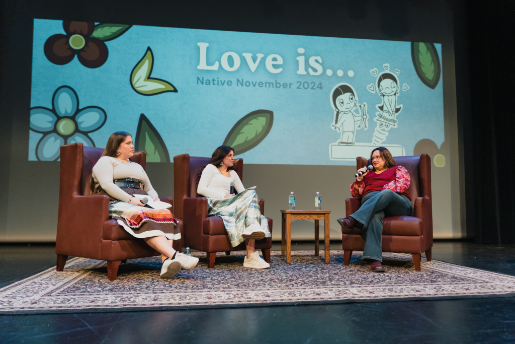 Two young women sit in chairs on a stage and laugh while a third women, also in a chair, makes a joke.