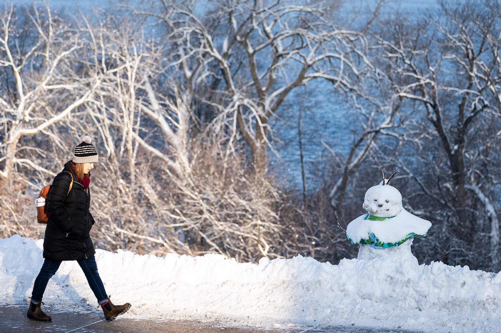 Person walking past a snowman wearing a scarf beside a snowy road with leafless, ice-covered trees in the background.