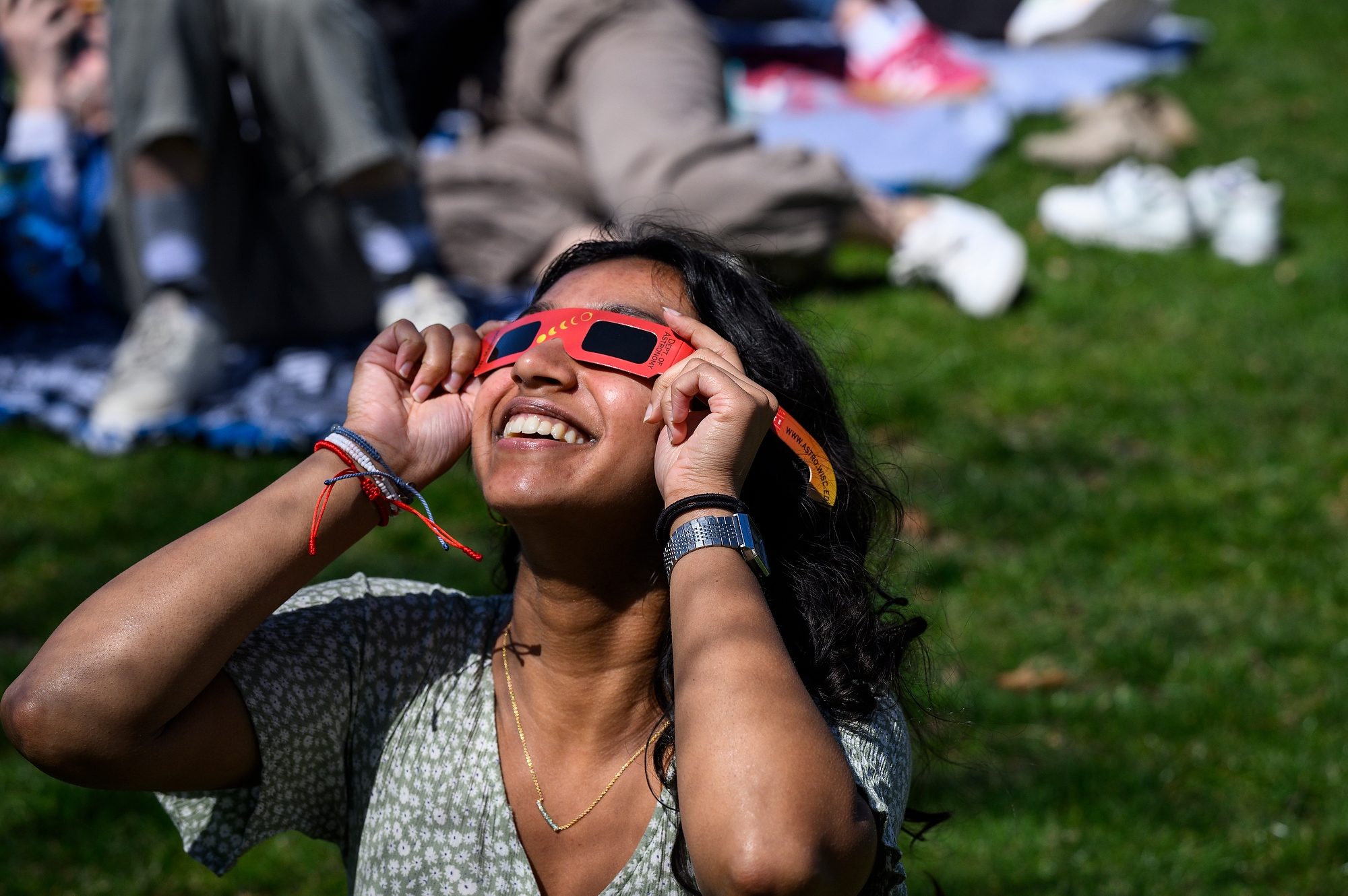 Person smiling and using eclipse glasses to look up to the sky in a sunny park setting.