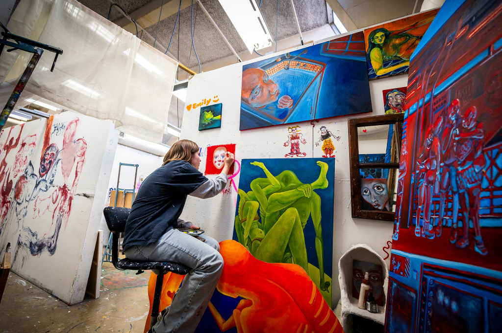 A woman carefully paints a face on a wall; the wall is hung with several other colorful paintings.