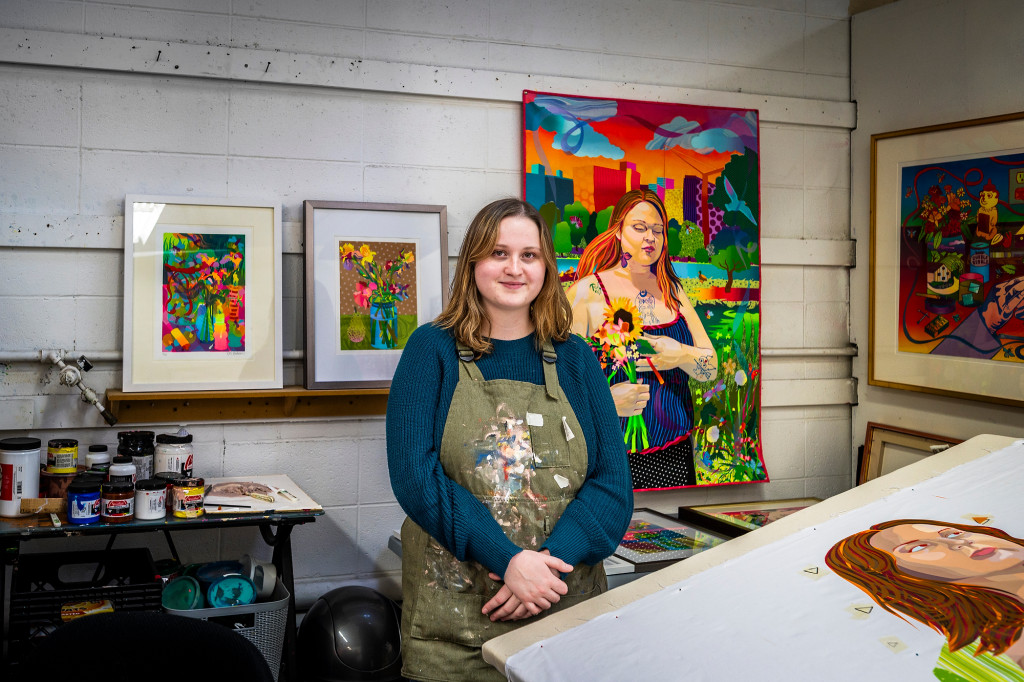 A woman stands in front of a wall hung with several very colorful artworks.