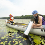 Two women in a canoe use equipment to take samples from the water.