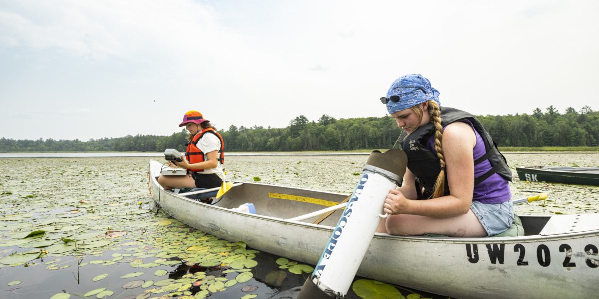 Two women in a canoe use equipment to take samples from the water.