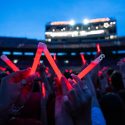 Audience members holding up glowing red sticks at a nighttime event in Camp Randall Stadium. In the foreground, someone holds the sticks to form a letter W.