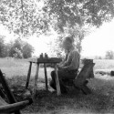 A black-and-white photo shows a man sitting at a table in an outdoors setting, writing in a notebook.