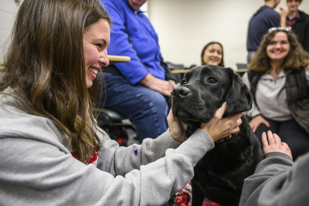 A woman smiles as she pets a black dog. Other people observe, smiling, in the background.