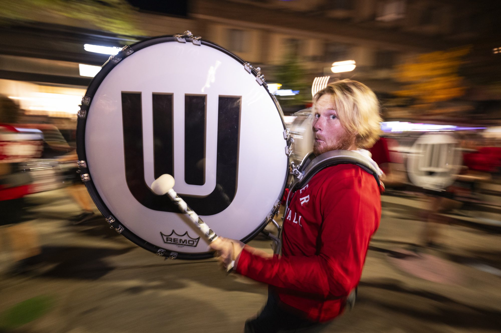 A person marching and playing a large bass drum with the University of Wisconsin logo on it during a nighttime parade.