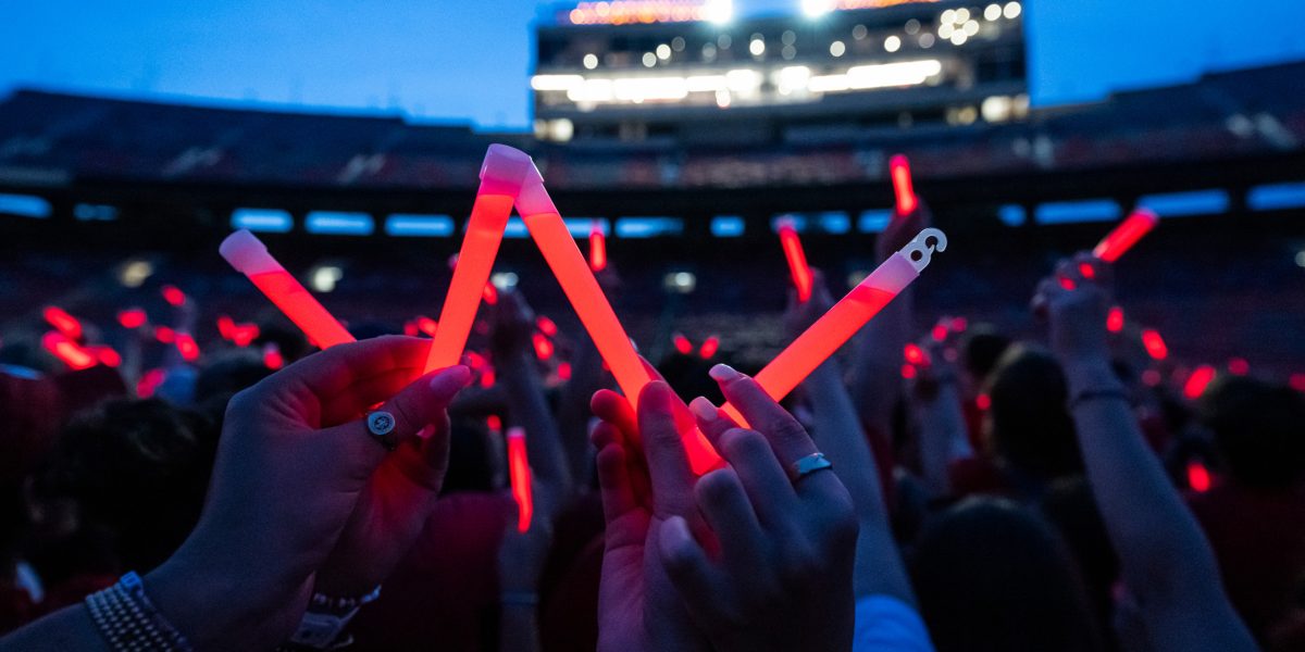 Audience members holding up glowing red sticks at a nighttime event in Camp Randall Stadium. In the foreground, someone holds the sticks to form a letter W.