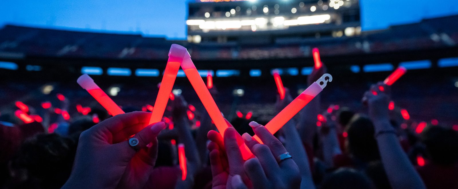 Audience members holding up glowing red sticks at a nighttime event in Camp Randall Stadium. In the foreground, someone holds the sticks to form a letter W.