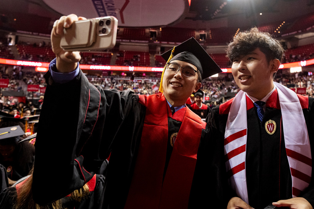 A person in a commencement gown takes a photo of himself and his friend.