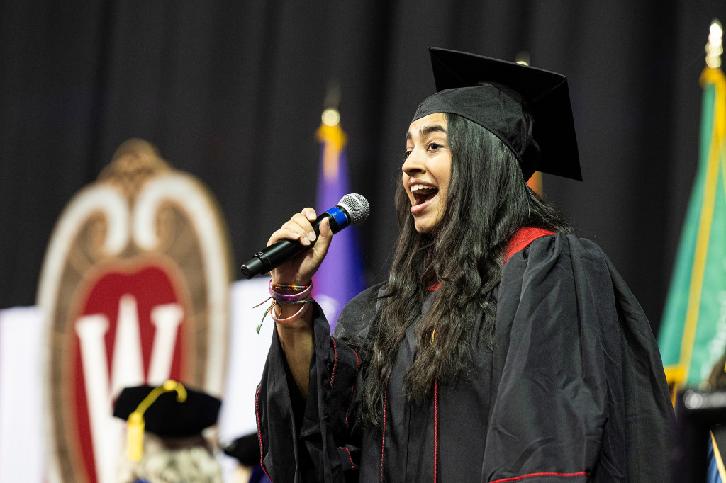 A woman sings on stage while wearing a commencement gown.