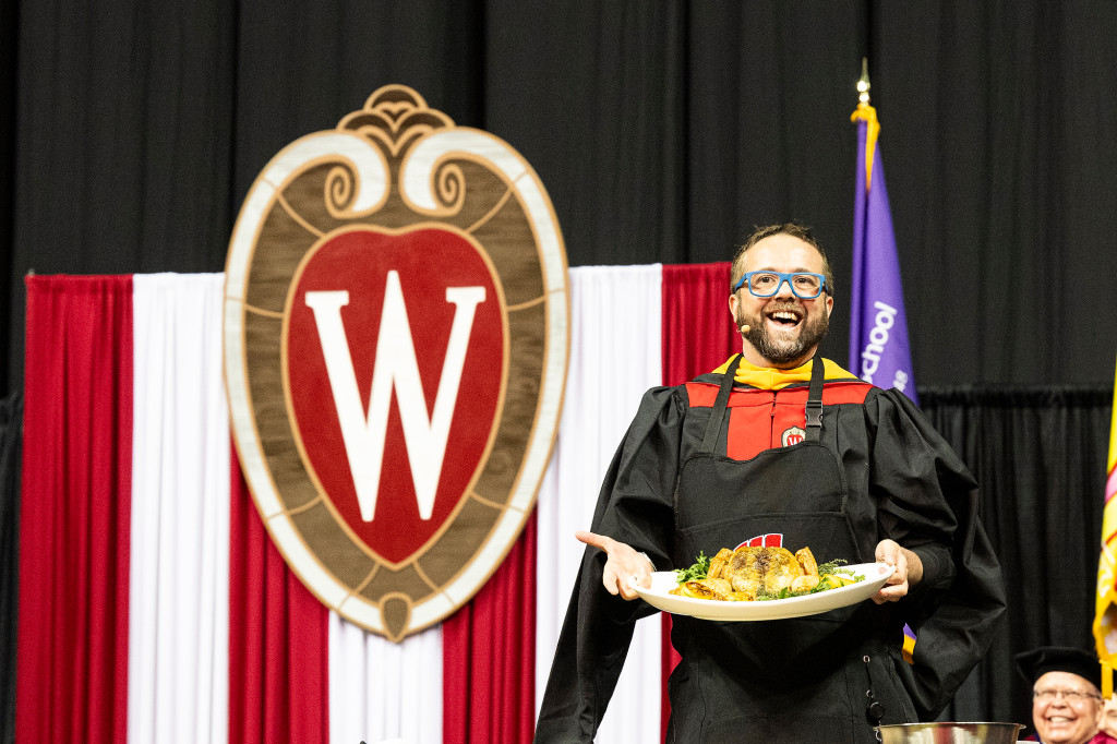 Photo of a man in academic robes holding up a roast chicken on a stage.