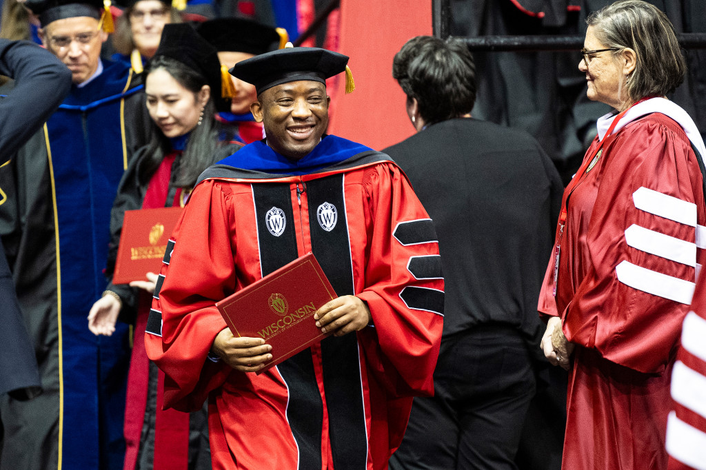 A man in a red academic robe holds a diploma cover and smiles.